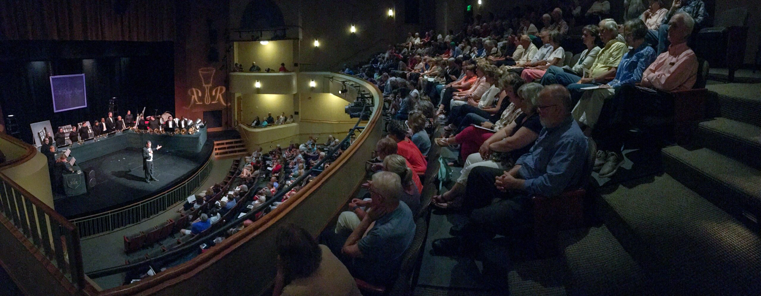 Members of The Raleigh Ringers and their director on stage in Hoover, Kansas. Shot includes members of the audience in the balcony and orchestra levels. The Raleigh ringers equipment is on display along with a projection of their logo on the wall.