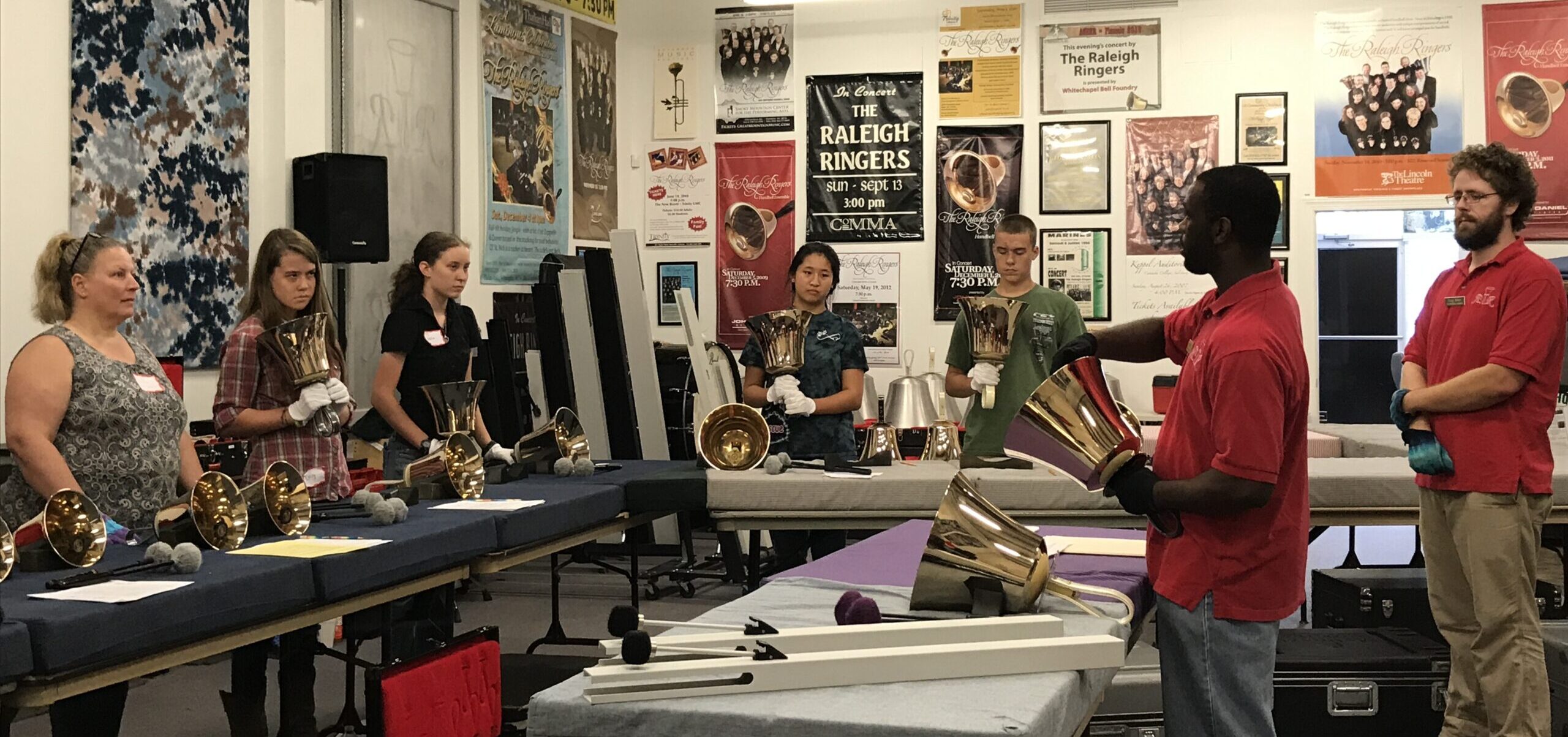 Five people standing in the rehearsal space for The Raleigh ringers being taught to ring handbells by two members of The Raleigh Ringers.