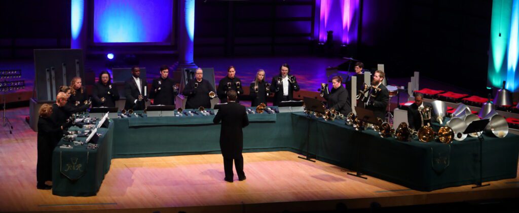 Members of The Raleigh Ringers performing at Meymandi Concert Hall in Raleigh, NC. 15 people stand behind tables with handbells and handchimes. In front of the tables is the director.