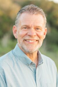 Photo of a man smiling into the camera wearing a blue collared shirt. The man is standing outside.